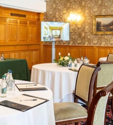 Elegant hotel meeting room with round tables, floral centrepieces, bottled water, notepads, and warm wood panelling.