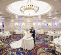 Elegant ballroom with grand chandelier, round tables, and a couple celebrating; ideal wedding venue at luxury hotel.