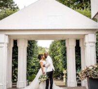 Elegant wedding couple embracing under a grand white pergola, surrounded by lush greenery and romantic floral arrangements.