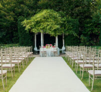 Elegant outdoor wedding venue with lush greenery, aisle seating, white floral arch, and romantic garden setting.